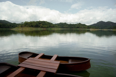 beautiful view of wooden boats on a lake with tree-lined hills under a cloudy blue skyの写真素材