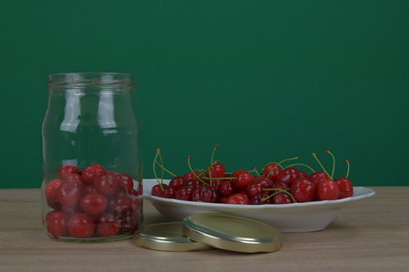 Ripe red cherries on a white plate on a wooden table. Homemade cherry jar. Green background.の写真素材