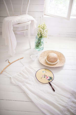 White summer dress and straw hat and handheld fan on the floor of white wooden room.の写真素材
