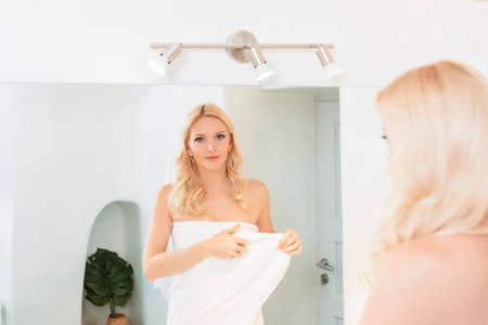 blond woman in a white bathroom looking at her reflection in the mirror, wearing a towel. High...