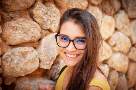 young woman standing in front of a stone wall. wearing glasses smiling at the camera. High quality photoの写真素材