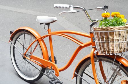 A bright orange bicycle with a wicker basket full of colorful yellow geraniums.  The bike has a matching white seat and white grips on the handlebars.の写真素材