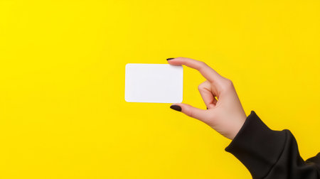 Woman hand with dark nail polish showing blank white card on yellow backgroundの素材