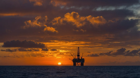 An offshore oil rig stands silhouetted against a vibrant sunset, casting reflections on the calm ocean. The sky is filled with dynamic clouds, creating a stunning backdrop.の素材