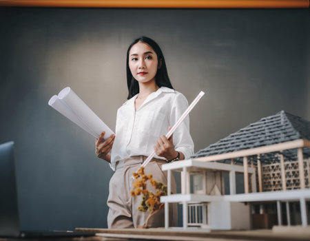 A confident architect stands in a contemporary office environment, holding blueprints in one hand while a detailed model of a house rests on the table beside her.の素材