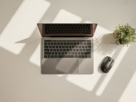 A laptop is open on a clean desk, accompanied by a wireless mouse and a small potted plant. Sunlight casts shadows, creating a warm and inviting workspace atmosphere.の素材