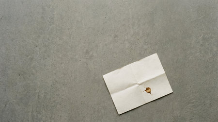 A crumpled piece of paper lays flat on a gray concrete surface. The paper shows a small stain, adding to its texture and visual interest in a minimalist setting.の素材
