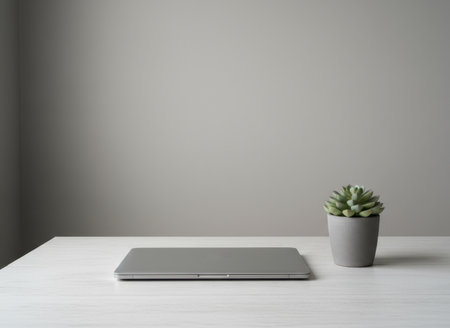 A laptop sits on a light wood table next to a small succulent plant in a gray pot. The plain wall in the background creates a simple workspace. This setup is suitable for productivity and focus.の素材