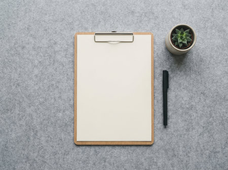 A blank clipboard sits on a gray surface next to a small plant and a pen. This setup is ideal for taking notes or drawing in a workspace that values simplicity.の素材