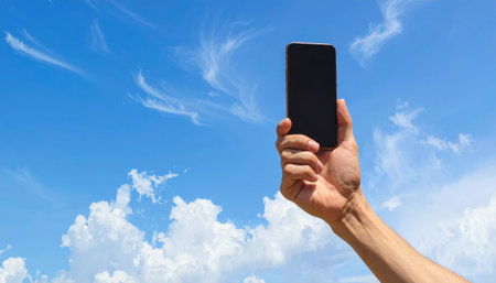 A person's arm and hand are raised, holding a dark smartphone up towards the sky, capturing the vast expanse of blue and scattered cloudsの素材