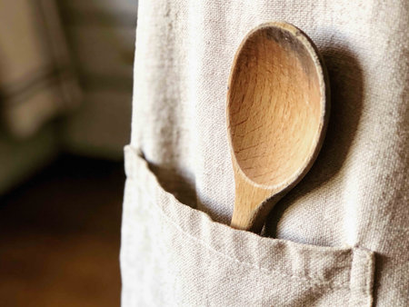 A close-up view of a well-used wooden spoon resting in the pocket of a textured linen apron, suggesting a home cooking environmentの素材