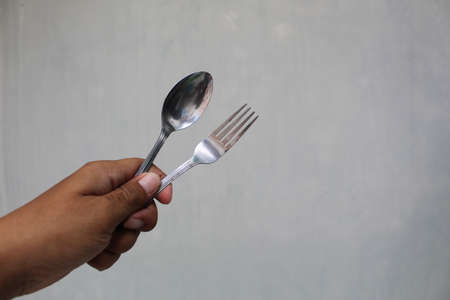 left hand holding a spoon and fork against a white wallの写真素材