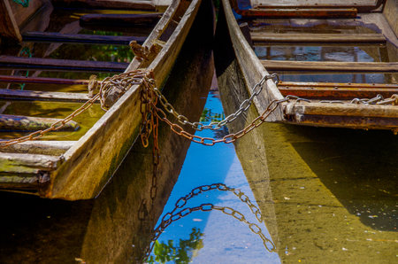 Two old wood barges connected with rusty chain. Chain is reflected in the waterの写真素材
