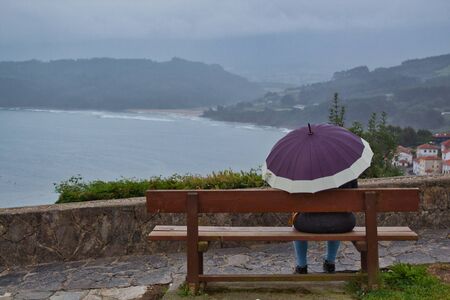Woman sitting on a bench in the rain with a purple umbrella on a cloudy and rainy day in from the sea.の写真素材