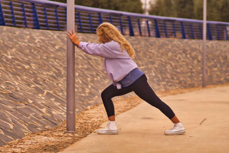 Young blonde woman performs stretching by a lamppost in a park ready to run, with a purple sweatshirt and black tights.の写真素材