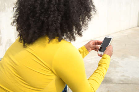 African american woman with afro hair and yellow t-shirt consulting her cell phone with her back turned.の写真素材