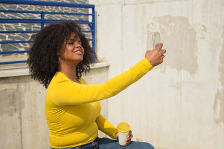 African american woman with afro hair holding a cup of coffee and wearing a yellow t-shirt taking a selfie with her cell phone sitting on a bench.の写真素材