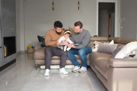 Gay married couple reading a story to their daughter on the sofa at home. Family diversity.の写真素材