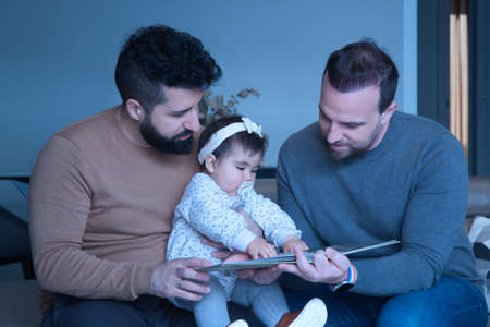 Gay married couple reading a story to their daughter on the sofa at home. Family diversity.の写真素材