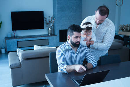Gay married couple chatting while one works at home the other holds their daughter in his arms in love. Family diversity.の写真素材
