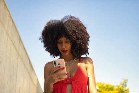 Young beautiful afro american woman with afro hair and coral top checking social networks on her cell phone.の写真素材