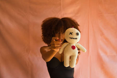 Young Latina woman in black dress holding voodoo doll in foreground on pink background.の写真素材