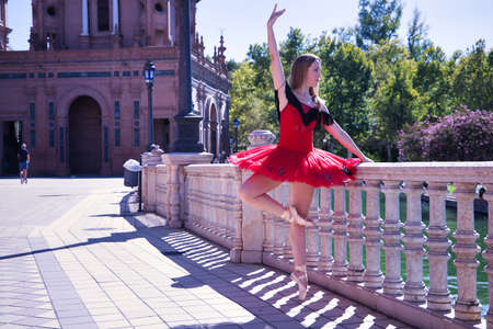 Adult female Hispanic classical ballet dancer in red tutu doing figures next to a stone railing in the middle of a plaza on a sunny day.の写真素材