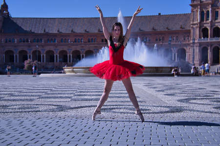 Hispanic adult female classical ballet dancer in red tutu doing figures in the middle of a plaza on a sunny day with a water fountain in the background.の写真素材