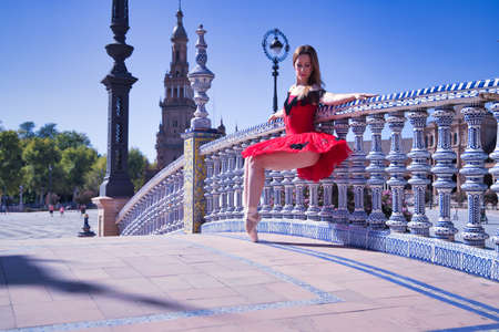 Adult female Hispanic classical ballet dancer in red tutu doing figures leaning on a beautiful tiled balustrade in the middle of a plaza on a sunny day.の写真素材