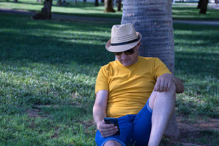 Mature adult Hispanic male wearing a hat and sunglasses, sitting on the grass and leaning against a tree while checking social networks on his cell phone.の写真素材