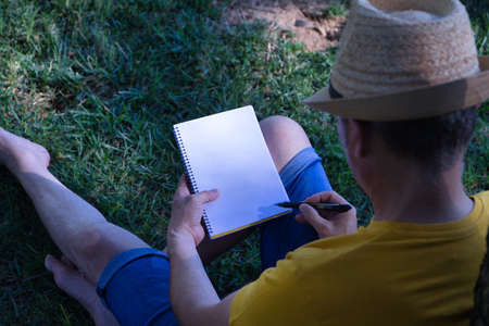 Hispanic mature adult man with a hat sitting on a park lawn and leaning on a tree with a blank notebook in one hand and a pen in the other.の写真素材