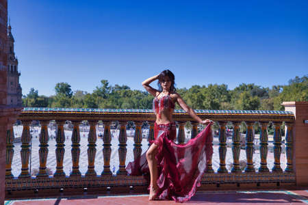 Middle-aged Hispanic woman, with red dress and rhinestones, to dance belly dance, making figures while dancing next to a tile railing. Belly dance concept.の写真素材