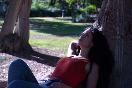 Middle-aged adult Hispanic woman with black curly hair, lying at the root of a tree, looking thoughtfully up at the sky. Concept thoughts, escape, nature.の写真素材