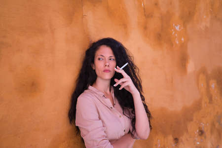 Middle-aged adult Hispanic woman with black curly hair, leaning against a yellow wall smoking. Concept smoking, cigar, tobacco, health.の写真素材