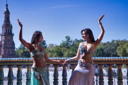 Two middle-aged Hispanic women, wearing beautifully colored dresses with rhinestones, to belly dance, making figures while dancing next to a tiled railing. Belly dance concept.の写真素材