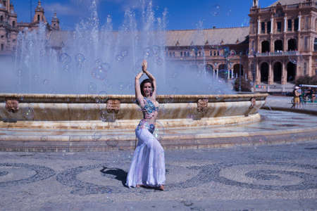Middle-aged Hispanic woman, wearing a beautifully colored dress with rhinestones, for belly dancing, dancing surrounded by soap bubbles in front of a water fountain. Belly dance concept.の写真素材