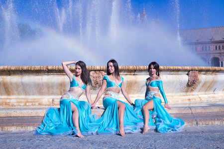 Three middle-aged Hispanic women, wearing turquoise costumes and rhinestones, to belly dance, sitting on a water fountain where a rainbow is formed. Belly dance concept.の写真素材
