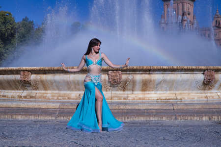 Middle-aged Hispanic woman, with turquoise costume and rhinestones, for belly dancing, dancing in front of a water fountain where a rainbow is formed. Belly dance concept.の写真素材