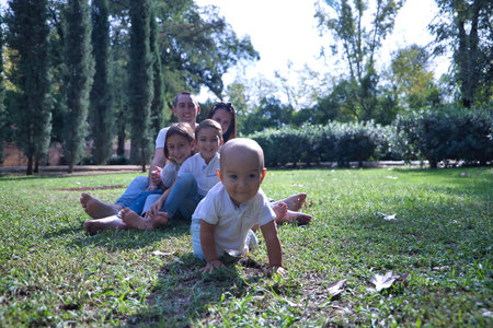 Happy, large family of five sitting on the lawn of a park, laughing as they watch the youngest one crawling away. Concept family, large family, happiness, first steps, crawling.の写真素材