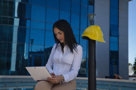 Young woman in white shirt, sitting, working with her tablet, with yellow worker's helmet and blueprint tube next to her. Business concept, architecture, construction, empowerment.の写真素材