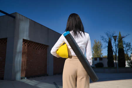 Young woman in white shirt, brown pants, with yellow workman's helmet under her arm and blueprint tube hanging on her back. Concept of business, architecture, construction, empowerment.の写真素材