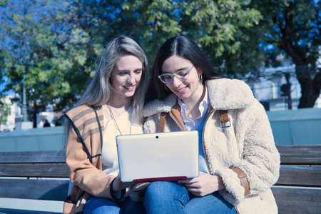 Portrait of real couple of young women, sitting on a bench consulting social networks on their laptop. Concept lgtbiq+, lesbians, tablet, technology.の写真素材