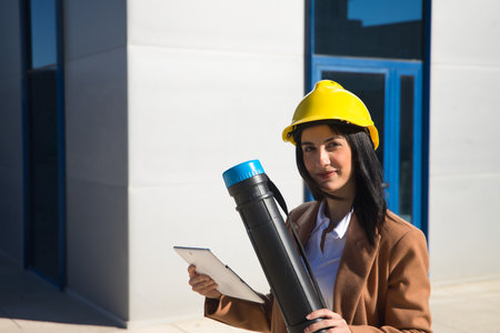 Young beautiful woman in brown coat, yellow workman's helmet and blueprint tube, working with her tablet. Business concept, architect, construction, working woman, empowerment.の写真素材