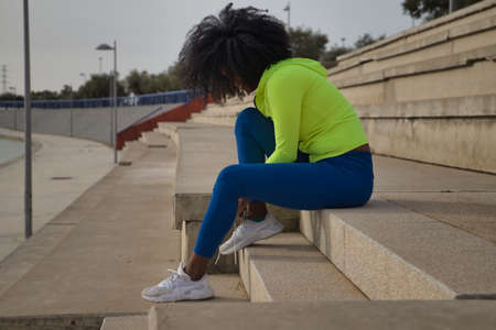 African-American woman with afro hair and sportswear, sitting, wearing leggings, buckling her laces to exercise in the street. Fitness concept, sport, street, urban.の写真素材
