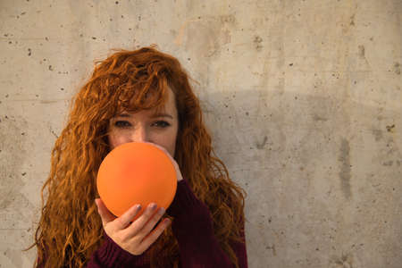 Portrait of young, attractive, red-haired, freckled, red-sweatered woman filling an orange globe on a gray wall. Concept colors, balloons, beauty, fashion, red.の写真素材