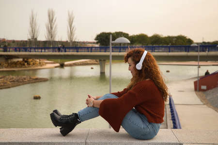 Young woman, red-haired, freckled, with red sweater and white headphones, sitting and concentrating, listening to music, in an outdoor park. Concept song, radio, music, playlist, mp3.の写真素材