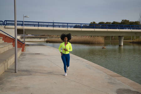 African-American woman with afro hair and sportswear, with fluorescent yellow sweatshirt and leggings, running through an outdoor park. Fitness concept, sport, street, urban, running.の写真素材