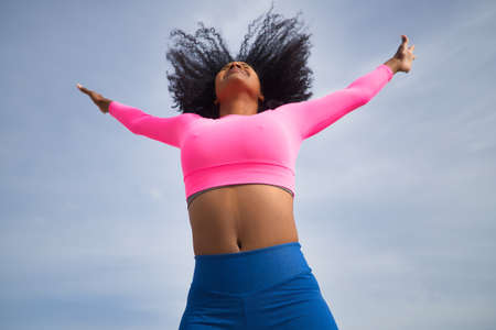 African-American woman with afro hair and sportswear, with fluorescent pink t-shirt and leggings, jumping into the sky, seen from below. Fitness concept, sport, street, urban, happiness.の写真素材