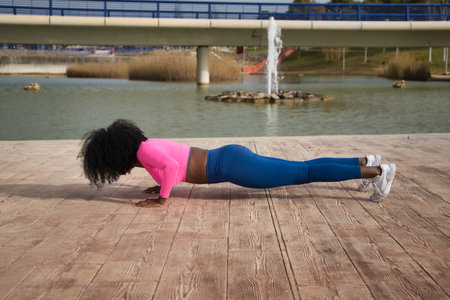 African-American woman with afro hair and sportswear, with fluorescent pink t-shirt and leggings, doing push-ups next to an outdoor lake. Fitness concept, sport, street, urban.の写真素材