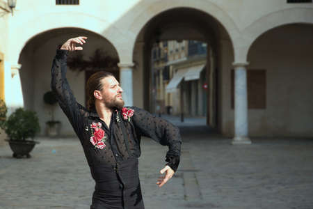 Young man with beard and long hair, wearing black transparent shirt with black polka dots and red roses, black pants and jacket, dancing flamenco in the city. Concept art, dance, culture, tradition.の写真素材